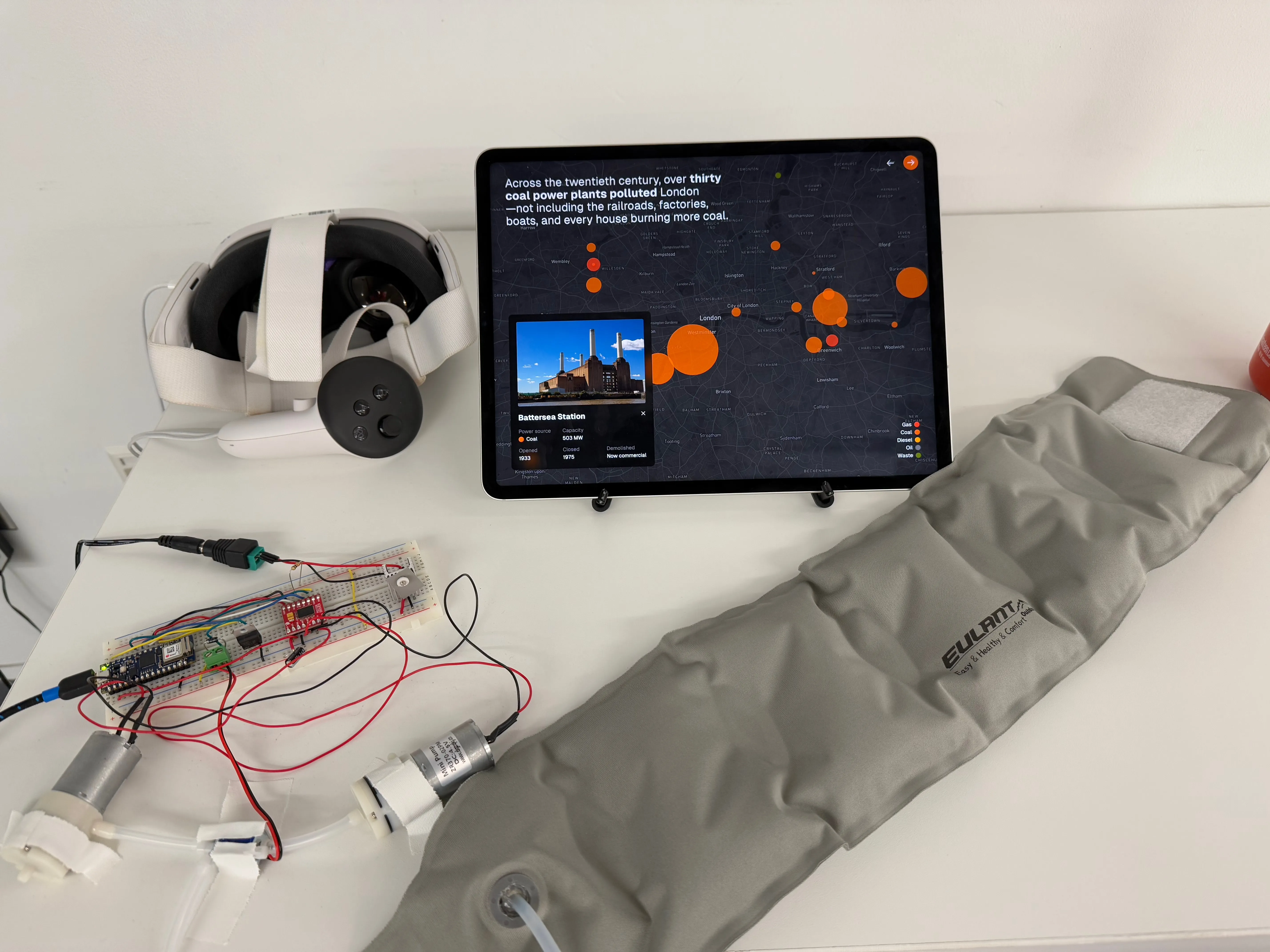 Grey neck pillow outstretched on a table with a breadboard and air pumps alongside a Meta Quest 3 VR headset and iPad showing a map of London